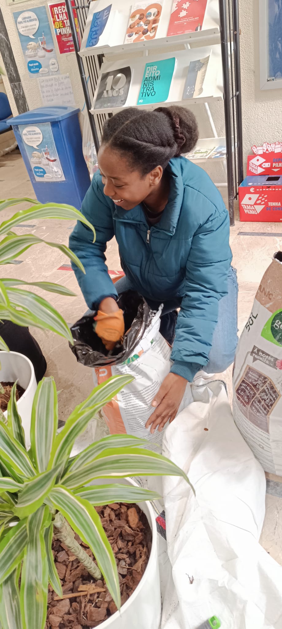 aluna com uma planta artificial a frente de uma estante com livros
